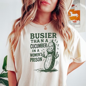 May include: Cream-colored t-shirt with the text "BUSIER THAN A CUCUMBER IN A WOMEN'S PRISON" and a cartoon cucumber illustration. The cucumber has a smiling face and thumbs up. The shirt is a casual style.