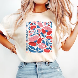 May include: Cream-colored t-shirt featuring a floral design in red, pink, and blue. The graphic includes the text "FREEDOM DEMOCRACY TRUTH" in a vertical column. The shirt is paired with light blue denim jeans.
