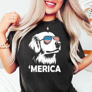 May include: Black t-shirt featuring a white illustration of a dog wearing sunglasses with an American flag design. The word 'MERICA is printed in white below the dog graphic. The shirt is a casual style.