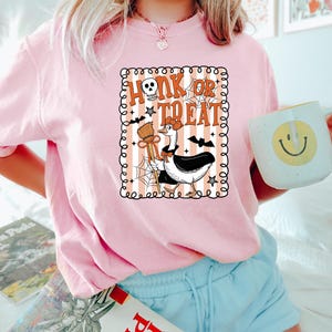 May include: Pink Halloween-themed t-shirt with the phrase "Honk or Treat" in orange and white. The graphic features a goose dressed as a witch with a broom and bat wings. The shirt is paired with blue shorts and a mug with a smiley face.