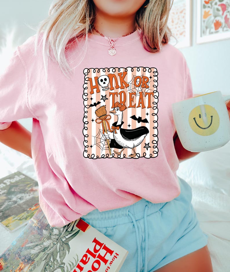 May include: Pink Halloween-themed t-shirt with the phrase "Honk or Treat" in orange and white. The graphic features a goose dressed as a witch with a broom and bat wings. The shirt is paired with blue shorts and a mug with a smiley face.