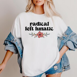 以下が含まれることがあります： 黒のスタイリッシュなフォントで「radical left lunatic」というテキストがプリントされた白いTシャツ。テキストの下には、赤い花と緑の葉のフローラルデザインがあります。Tシャツはデニムジャケットと合わせて着用されています。