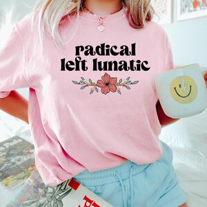 以下が含まれることがあります： 「radical left lunatic」の黒い文字と花の模様が入った、薄いピンク色のTシャツ。黄色いスマイリーフェイスの白いマグカップを持っています。