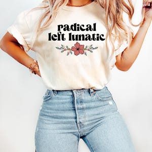 以下が含まれることがあります： 「radical left lunatic」の黒い文字と花の模様がプリントされたクリーム色のTシャツ。明るいブルーデニムのジーンズと組み合わせています。全体的にカジュアルでリラックスしたスタイルです。