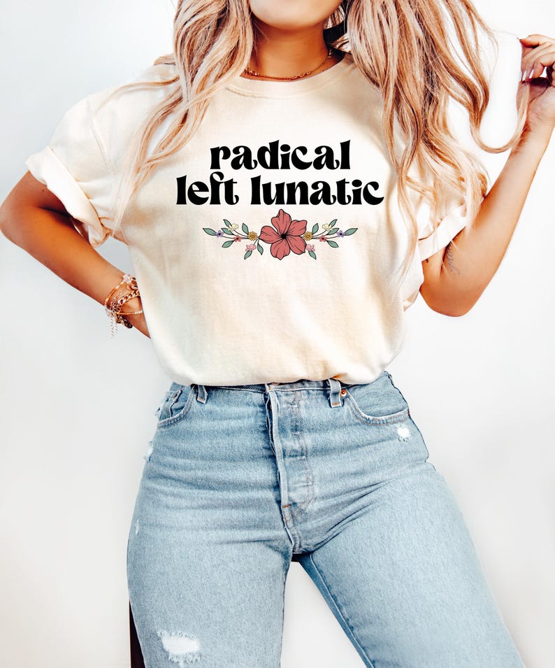 以下が含まれることがあります： 「radical left lunatic」の黒い文字と花の模様がプリントされたクリーム色のTシャツ。明るいブルーデニムのジーンズと組み合わせています。全体的にカジュアルでリラックスしたスタイルです。