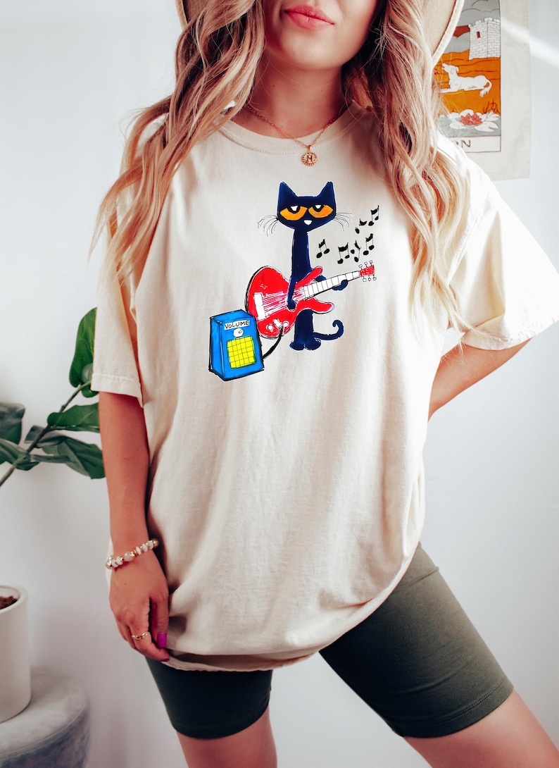 May include: Cream-colored t-shirt featuring a cartoon black cat playing a red guitar. The cat has yellow eyes and is surrounded by musical notes. A blue amplifier with the word "VOLUME" is next to the cat. The shirt is a casual, relaxed fit.