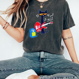 May include: Dark gray t-shirt featuring a cartoon cat playing a red guitar. The cat is black with yellow eyes and is surrounded by musical notes. A blue amplifier with a yellow speaker is next to the cat.