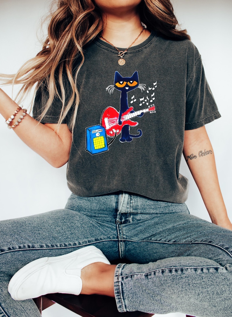 May include: Dark gray t-shirt featuring a cartoon cat playing a red guitar. The cat is black with yellow eyes and is surrounded by musical notes. A blue amplifier with a yellow speaker is next to the cat.
