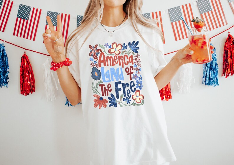 May include: White t-shirt with the text "America Land of The Free" in red, blue, and green floral design. The image is decorated with American flags and red, white, and blue decorations. A person is holding a drink with a strawberry.