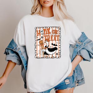 May include: White t-shirt featuring a Halloween-themed graphic. The design includes the text "Honk or Treat" with a skull, a goose dressed in a witch costume, bats, stars, and a broom. The graphic is framed with a decorative border.