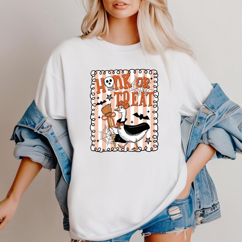 May include: White t-shirt featuring a Halloween-themed graphic. The design includes the text "Honk or Treat" with a skull, a goose dressed in a witch costume, bats, stars, and a broom. The graphic is framed with a decorative border.
