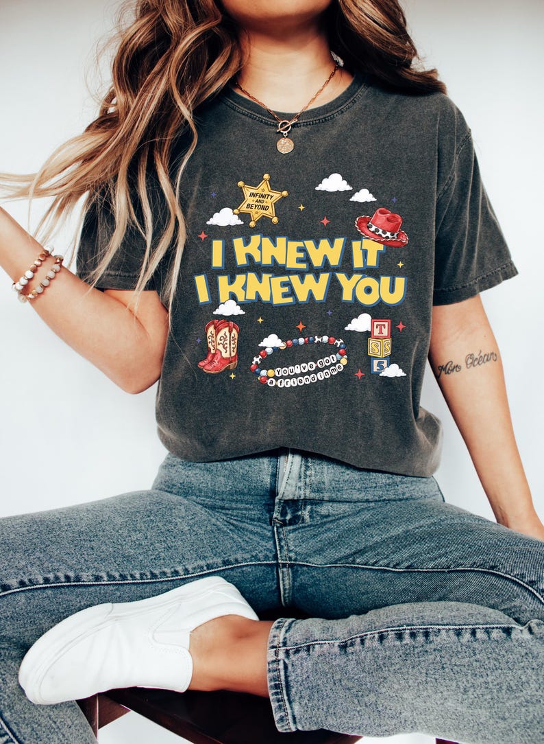 May include: A charcoal gray short-sleeve t-shirt featuring a graphic print with the text I KNEW IT I KNEW YOU. The design includes a yellow sheriff star, a red cowboy hat, red cowboy boots, a beaded bracelet, and alphabet blocks with clouds.