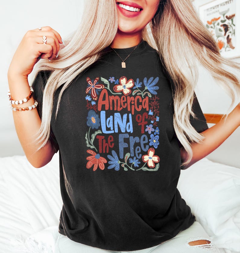 May include: Black t-shirt with the text "America Land of The Free" in a floral design. The design features red, blue, and white flowers and leaves. The shirt is a dark color and has a relaxed fit.