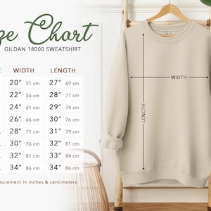 Puede incluir: Una sudadera beige Gildan 18000 cuelga de un estante de madera, con medidas de ancho y largo. Una tabla de tallas muestra las tallas S a 5XL, con medidas en pulgadas y cent&iacute;metros. La tabla incluye un ancho de 51 cm a 86 cm y un largo de 69 cm a 86 cm.