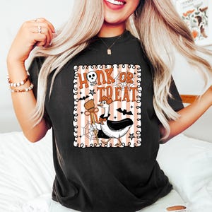May include: Black t-shirt with a Halloween-themed graphic. The graphic features a cartoon goose dressed in a witch's hat, holding a broom, with the words "Honk or Treat" in a spooky font. Bats, stars, and spiderwebs complete the design.
