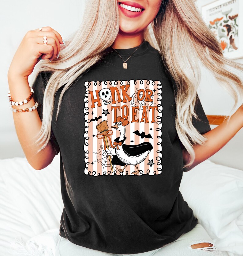 May include: Black t-shirt with a Halloween-themed graphic. The graphic features a cartoon goose dressed in a witch's hat, holding a broom, with the words "Honk or Treat" in a spooky font. Bats, stars, and spiderwebs complete the design.