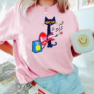May include: Pink t-shirt featuring a black cat playing a red guitar, with musical notes and a blue amplifier. The cat has yellow eyes and is wearing a black bow tie. A white mug with a yellow smiley face is held in the hand.