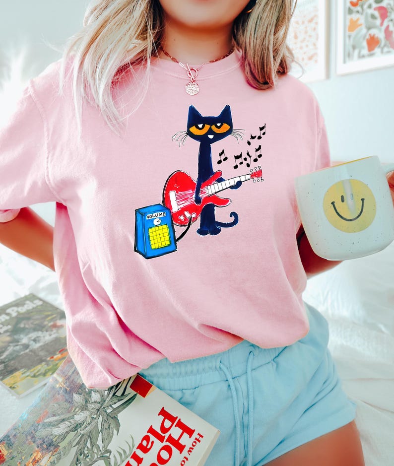 May include: Pink t-shirt featuring a black cat playing a red guitar, with musical notes and a blue amplifier. The cat has yellow eyes and is wearing a black bow tie. A white mug with a yellow smiley face is held in the hand.