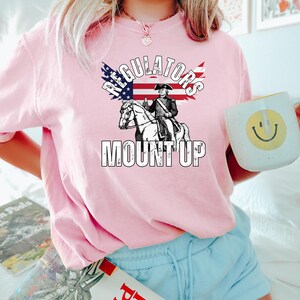 May include: Pale pink sweatshirt with the text "Regulators Mount Up" and a graphic of a figure on horseback, with an American flag design. The sweatshirt is a light pink colour.