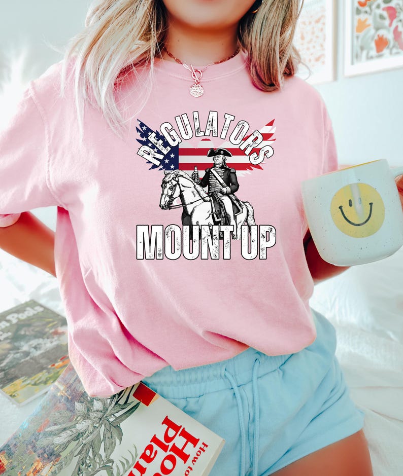 May include: Pale pink sweatshirt with the text "Regulators Mount Up" and a graphic of a figure on horseback, with an American flag design. The sweatshirt is a light pink colour.