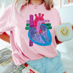 Puede incluir: Una camiseta rosa claro con un diagrama anat&oacute;mico del coraz&oacute;n colorido con etiquetas como "aorta" y "ventr&iacute;culo". El gr&aacute;fico es en tonos de rojo, azul y morado. La persona sostiene una taza blanca con una carita sonriente amarilla.