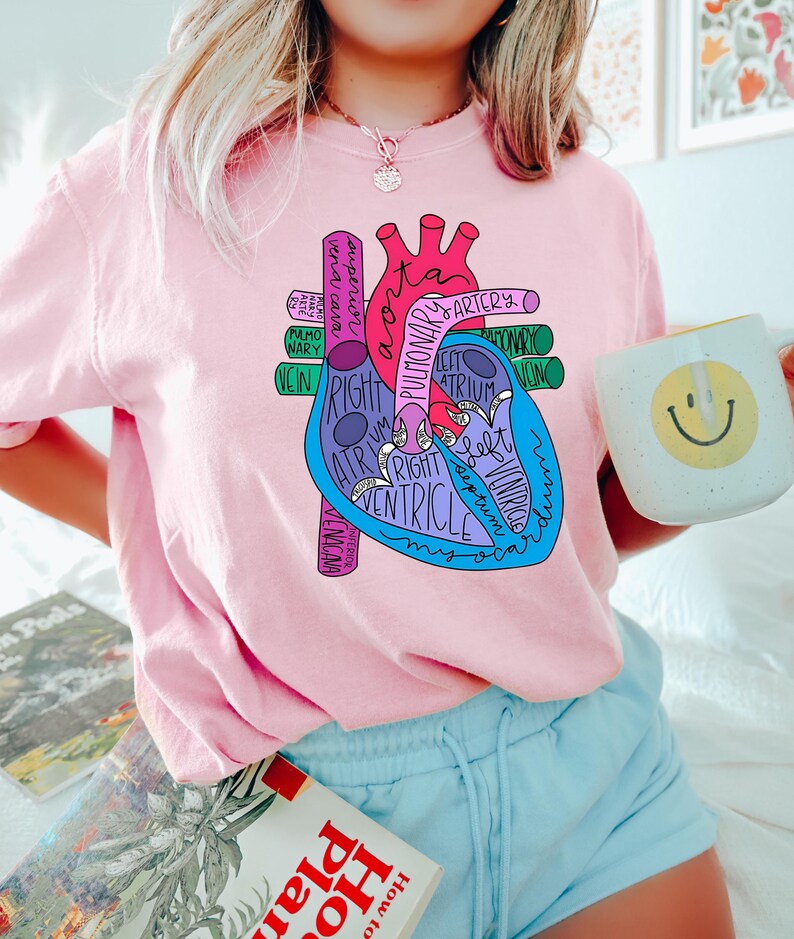 Puede incluir: Una camiseta rosa claro con un diagrama anat&oacute;mico del coraz&oacute;n colorido con etiquetas como "aorta" y "ventr&iacute;culo". El gr&aacute;fico es en tonos de rojo, azul y morado. La persona sostiene una taza blanca con una carita sonriente amarilla.