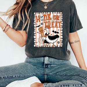 May include: Dark gray t-shirt with a Halloween-themed graphic. The graphic features a goose dressed as a witch with the words "Honk or Treat" in orange and white. The design includes bats, a skull, and a broom.
