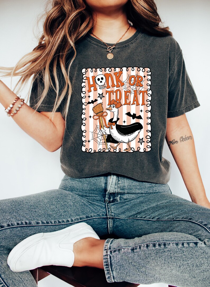 May include: Dark gray t-shirt with a Halloween-themed graphic. The graphic features a goose dressed as a witch with the words "Honk or Treat" in orange and white. The design includes bats, a skull, and a broom.