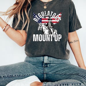 May include: Dark grey t-shirt with the words "REGULATORS MOUNT UP" in white. The graphic features an illustration of a man on a horse with an American flag design. A casual wear item.
