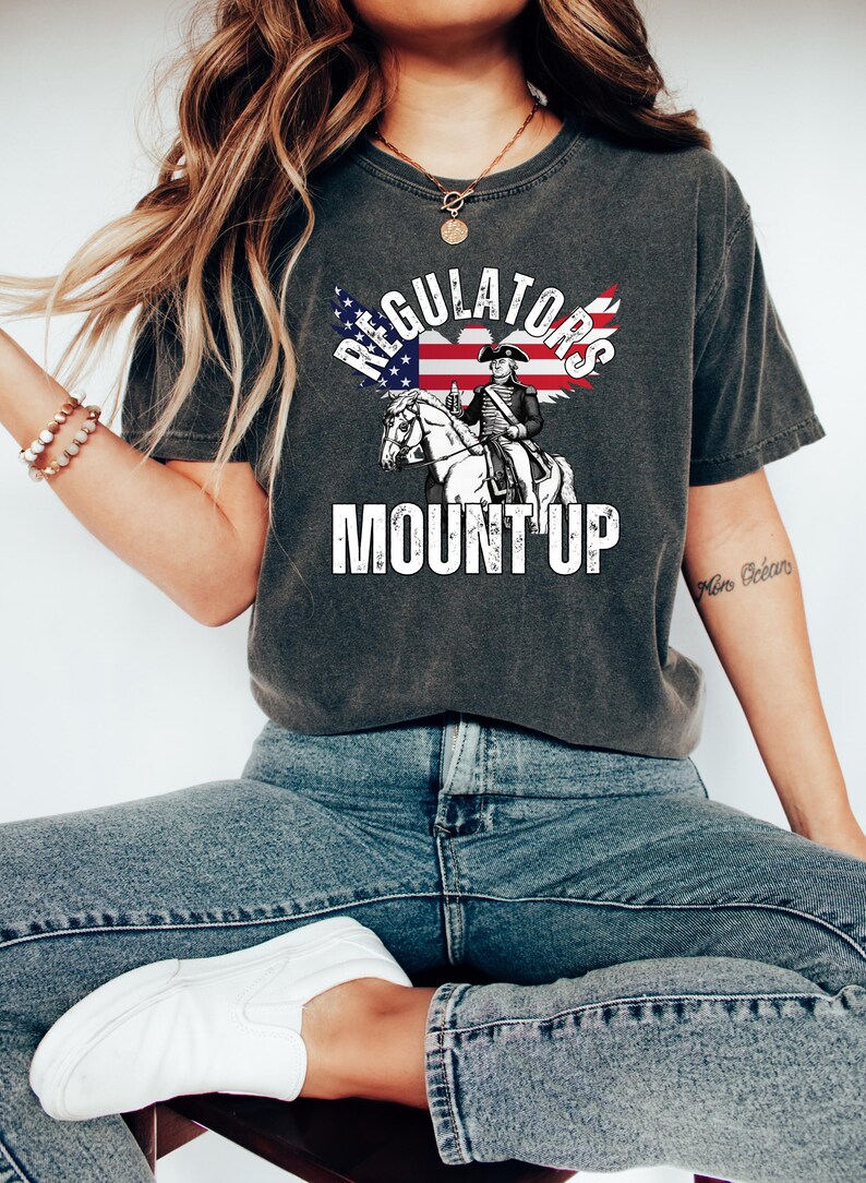 May include: Dark grey t-shirt with the words "REGULATORS MOUNT UP" in white. The graphic features an illustration of a man on a horse with an American flag design. A casual wear item.