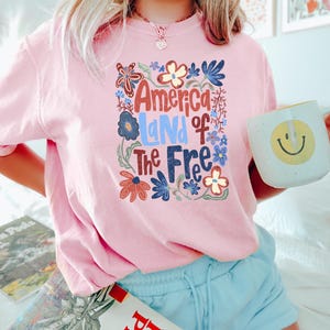 May include: Pink t-shirt with the text "America Land of the Free" in a floral design. The shirt is paired with light blue shorts. A white mug with a yellow smiley face is held in the hand.