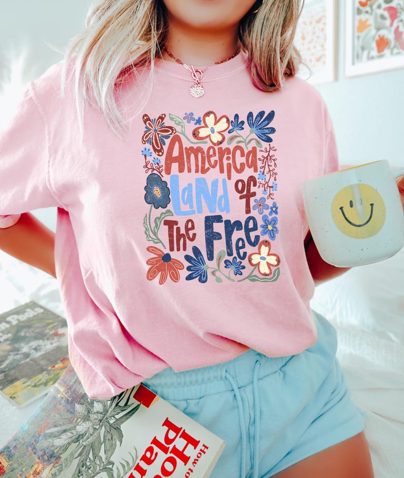 May include: Pink t-shirt with the text "America Land of the Free" in a floral design. The shirt is paired with light blue shorts. A white mug with a yellow smiley face is held in the hand.
