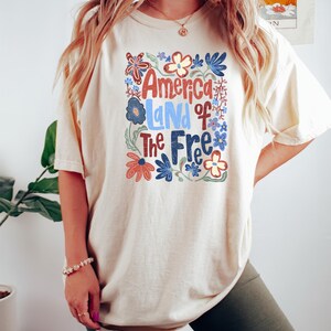 May include: Cream-colored t-shirt with the text "America Land of The Free" in red, blue, and white, surrounded by floral designs. The shirt is a casual style, suitable for everyday wear.
