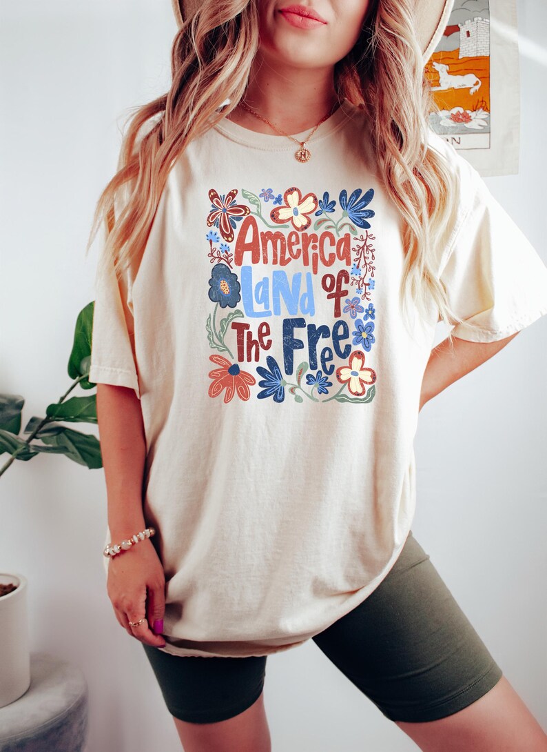 May include: Cream-colored t-shirt with the text "America Land of The Free" in red, blue, and white, surrounded by floral designs. The shirt is a casual style, suitable for everyday wear.