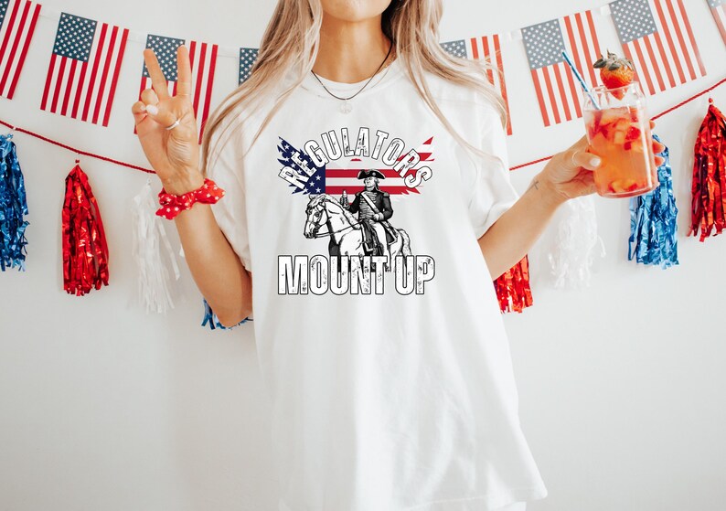 May include: White t-shirt with a patriotic design featuring an illustration of a man on a horse, the American flag, and the words "REGULATORS MOUNT UP." The shirt is decorated with red, white, and blue accents, and American flags are in the background.