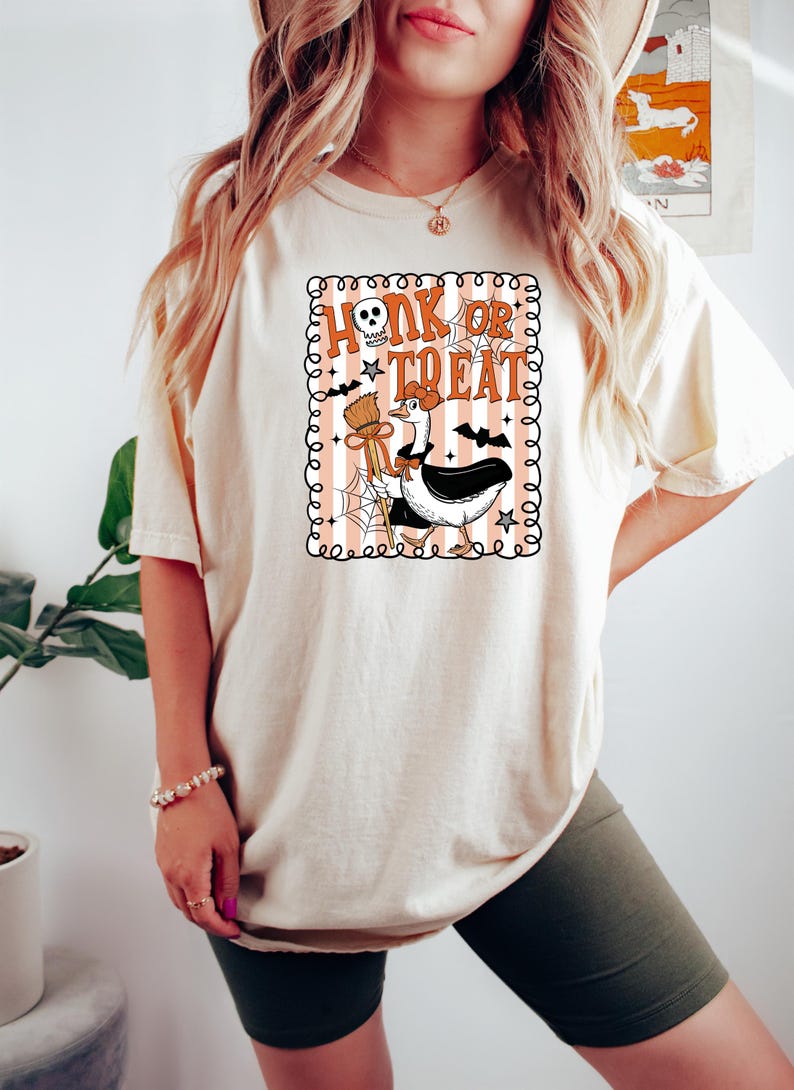 May include: Cream-colored t-shirt with a Halloween-themed graphic. The graphic features a goose dressed as a witch, holding a broom and a spider web, with the text "Honk or Treat" in orange and black.