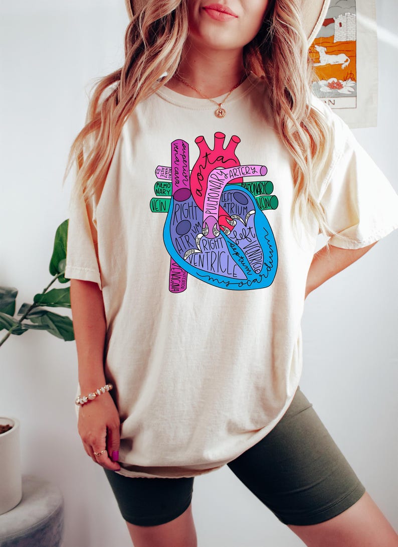 Puede incluir: Camiseta color crema con un colorido diagrama anat&oacute;mico del coraz&oacute;n. El diagrama incluye etiquetas como "aorta", "arteria pulmonar" y "ventr&iacute;culo derecho". El dise&ntilde;o es en tonos de rosa, azul y morado.
