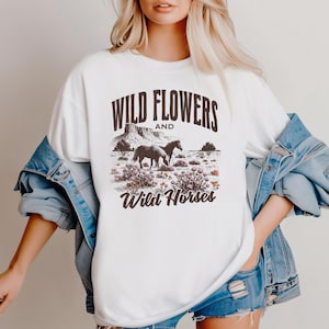 May include: White t-shirt with a graphic design featuring the text "Wild Flowers and Wild Horses." The design includes illustrations of horses, desert plants, and a mountain landscape in shades of brown and green.