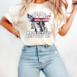 May include: A cream-coloured t-shirt with a graphic of a man on horseback, an American flag, and the words "REGULATORS" and "MOUNT UP". The design is in black and white with red and blue accents.