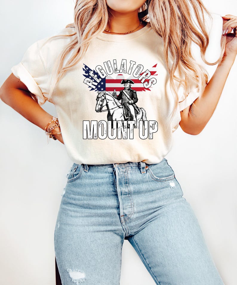 May include: A cream-coloured t-shirt with a graphic of a man on horseback, an American flag, and the words "REGULATORS" and "MOUNT UP". The design is in black and white with red and blue accents.