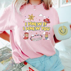 May include: A light pink t-shirt featuring a graphic print with the text I KNEW IT I KNEW YOU, a yellow sheriff star badge, a red cowboy hat, a pair of cowboy boots, a beaded bracelet with the text YOU'VE GOT A FRIEND IN ME, and toy blocks.