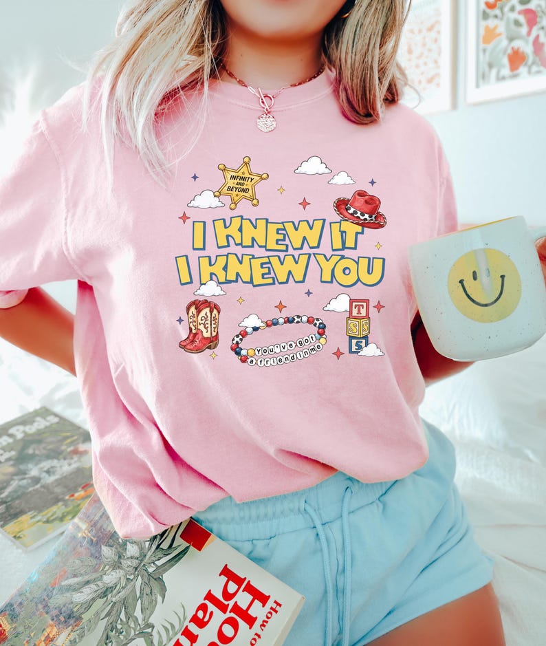 May include: A light pink t-shirt featuring a graphic print with the text I KNEW IT I KNEW YOU, a yellow sheriff star badge, a red cowboy hat, a pair of cowboy boots, a beaded bracelet with the text YOU'VE GOT A FRIEND IN ME, and toy blocks.