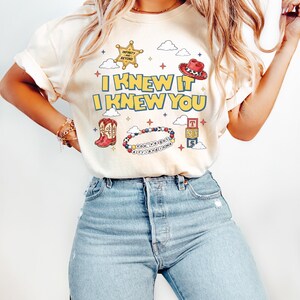 May include: A cream-colored t-shirt featuring the yellow text I KNEW IT I KNEW YOU. The graphic includes a gold sheriff badge, a red cowboy hat, red cowboy boots, a beaded bracelet with the text You've got a friend in me, and toy blocks.