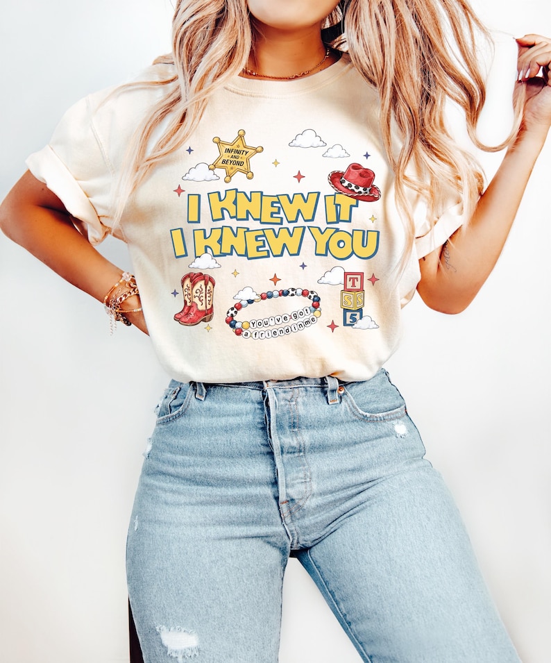 May include: A cream-colored t-shirt featuring the yellow text I KNEW IT I KNEW YOU. The graphic includes a gold sheriff badge, a red cowboy hat, red cowboy boots, a beaded bracelet with the text You've got a friend in me, and toy blocks.