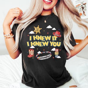 May include: A black t-shirt featuring a graphic print with the yellow text I KNEW IT I KNEW YOU. The design includes a gold sheriff badge with the text INFINITY AND BEYOND, a red cowboy hat, red cowboy boots, a beaded bracelet with the text YOU'VE GOT A FRIEND IN ME, and three stacked alphabet blocks with the letters T, S, and S.