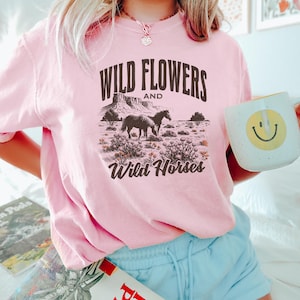 May include: Pink t-shirt with a graphic that reads "Wild Flowers and Wild Horses." The graphic depicts two horses in a desert landscape. The person is holding a white mug with a yellow smiley face.