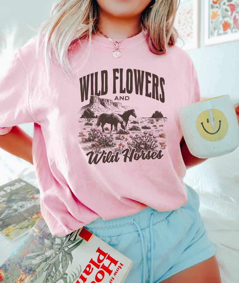May include: Pink t-shirt with a graphic that reads "Wild Flowers and Wild Horses." The graphic depicts two horses in a desert landscape. The person is holding a white mug with a yellow smiley face.