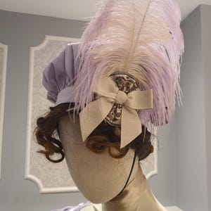May include: A lavender-colored hat with a large, feathery plume. The hat features a decorative bow and a circular embellishment. The hat is part of a costume or historical fashion ensemble.