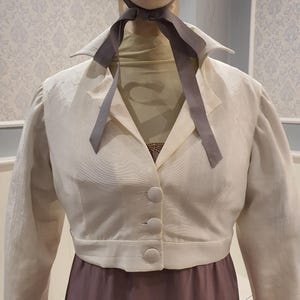 May include: Cream-colored, button-down jacket with a notched collar and long sleeves. The jacket is paired with a mauve-colored dress. A gray ribbon is tied around the neck of a mannequin. The outfit is displayed on a mannequin.