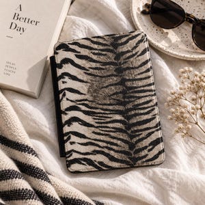 May include: A rectangular e-reader case featuring a black and beige tiger stripe pattern. The case has a smooth, textured surface and a slim profile, designed to protect a digital device. The cover is closed, displaying the animal print design.